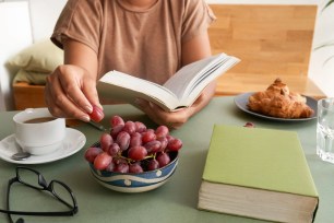 Unrecognizable woman wrapped up in reading novel while sitting at kitchen table and eating ripe grape, cup of fragrant coffee and appetizing croissants served for breakfast